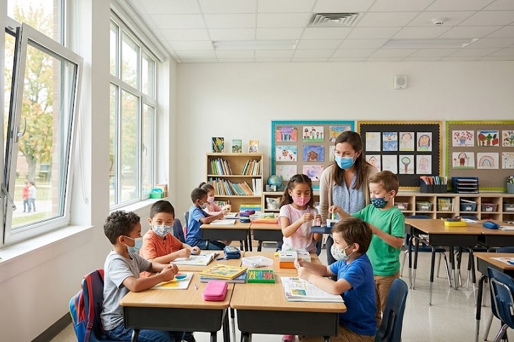 A bright, well-ventilated elementary classroom where young students wearing colorful face masks work at their desks while a teacher in a medical mask assists them.