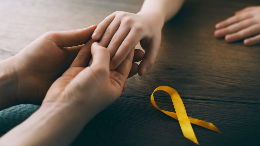 Hands holding together in a supportive gesture beside a yellow cancer awareness ribbon, representing pediatric cancer survivorship and compassionate care.