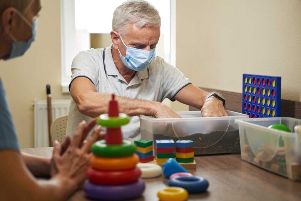 A therapist helps a patient with hand exercises during the stroke recovery process. The patient uses colorful stackable rings and a container with sensory material.