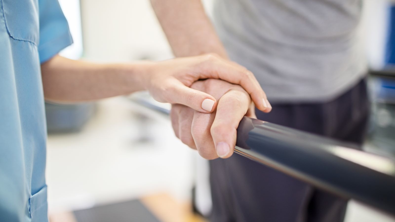 Close-up of a patient's hand on a parallel bar during the stroke recovery process. A clinician in blue scrubs guides the patient's movement.