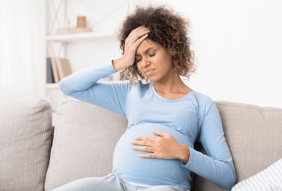 Pregnant woman holding her forehead in pain while sitting on a couch, showing headache symptoms managed by low-dose aspirin during pregnancy.
