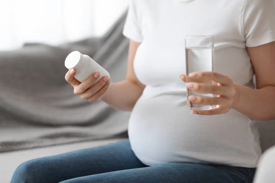Pregnant woman sitting on a sofa holding a medicine bottle and a glass of water to safely take low-dose aspirin during pregnancy.