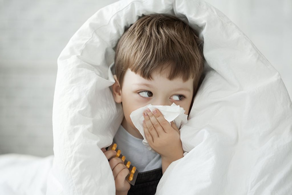 Young boy wrapped in a blanket blowing his nose with a tissue while holding medicine during cold and flu season.