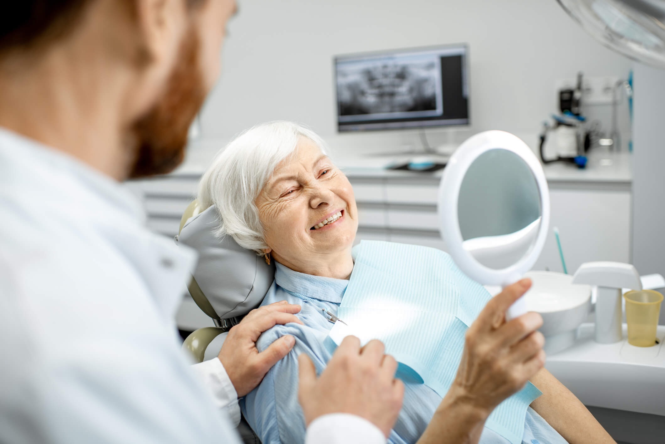 An old female patient with happy white teeth looking into a white hand-held mirror, promoting healthy teeth for life.