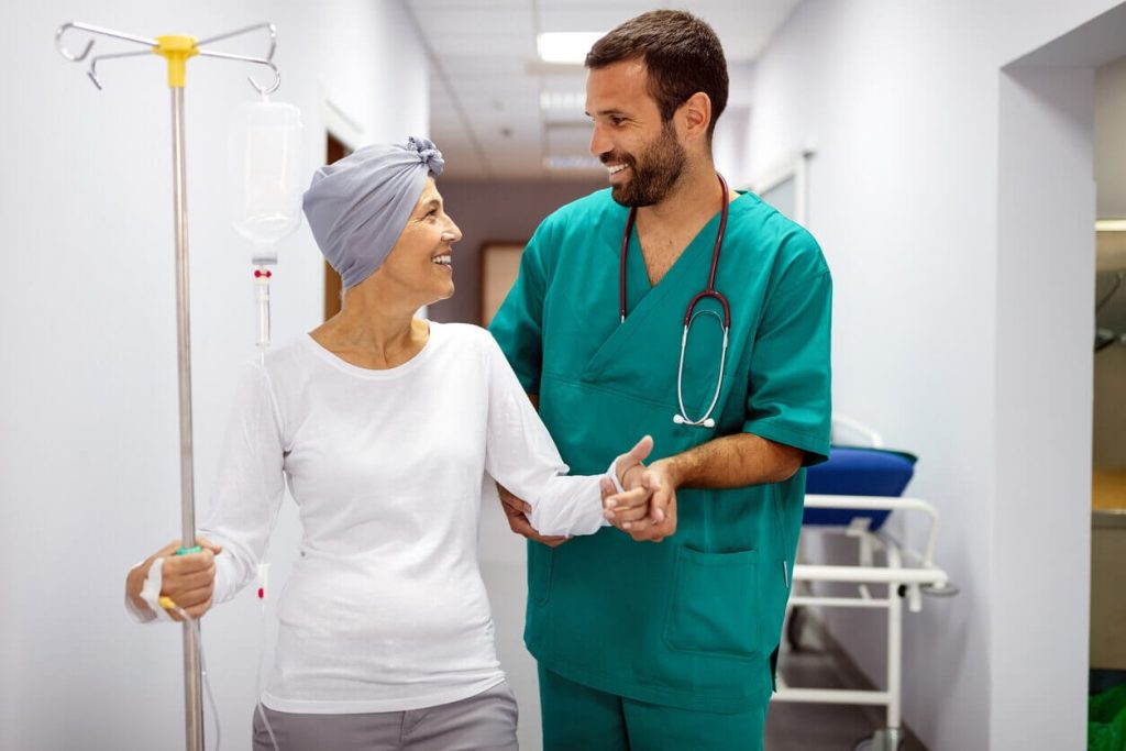 A male doctor assisting a smiling female patient in a headscarf along a clinic hallway, supporting local cancer care access.