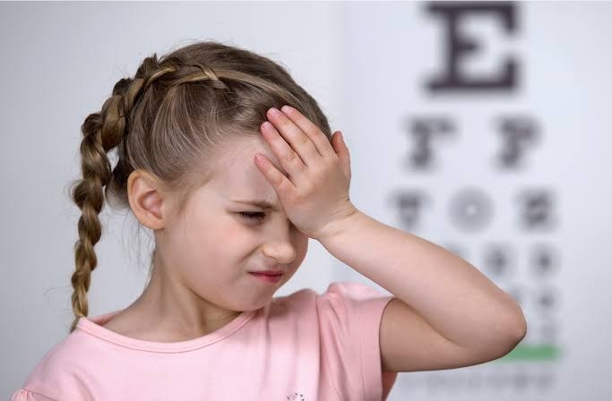 A young girl squinting and holding her forehead in front of an eye chart, highlighting signs of myopia in children.